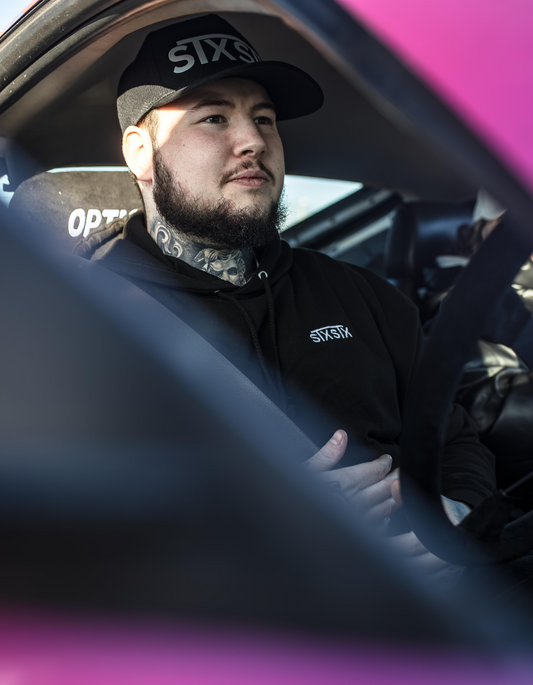 A person wearing a black snapback hat with the brand name 'STXSTX' visible on the front, sitting in a car with pink interior.
