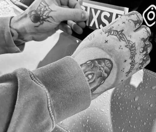 A close-up black and white image of a person's hand with a tattoo, holding a small sticker with the text 'STXSTX'.