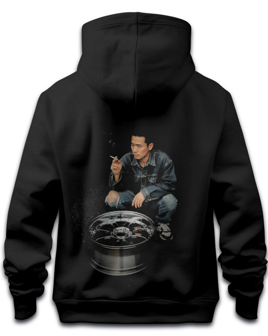 Black hoodie with a graphic of a japanese drifter person crouched over an alloy wheel expressing japanese drift culture on the back, on a white background