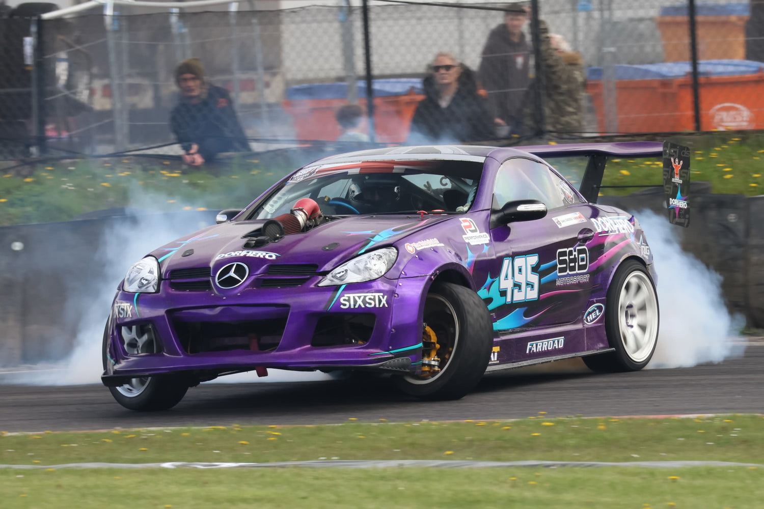 Purple Mercedes-Benz car performing a drift on a track with spectators in the background.