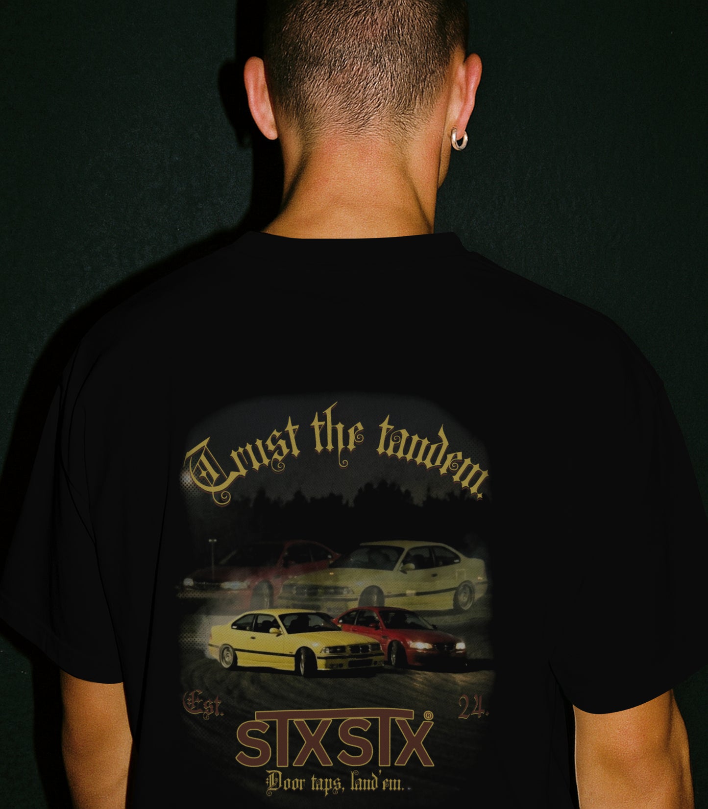 rear view of a male model wearing a STXSTX premium Black Tshirt with a rear design of 2 bmws in tandem drift on a racetrack