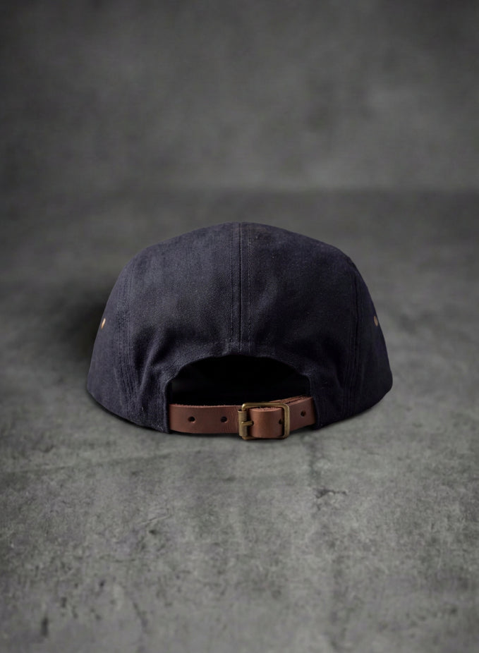 A dark black canvas baseball cap viewed from the back, featuring a brown leather strap and brass buckle closure, set against a textured gray background.