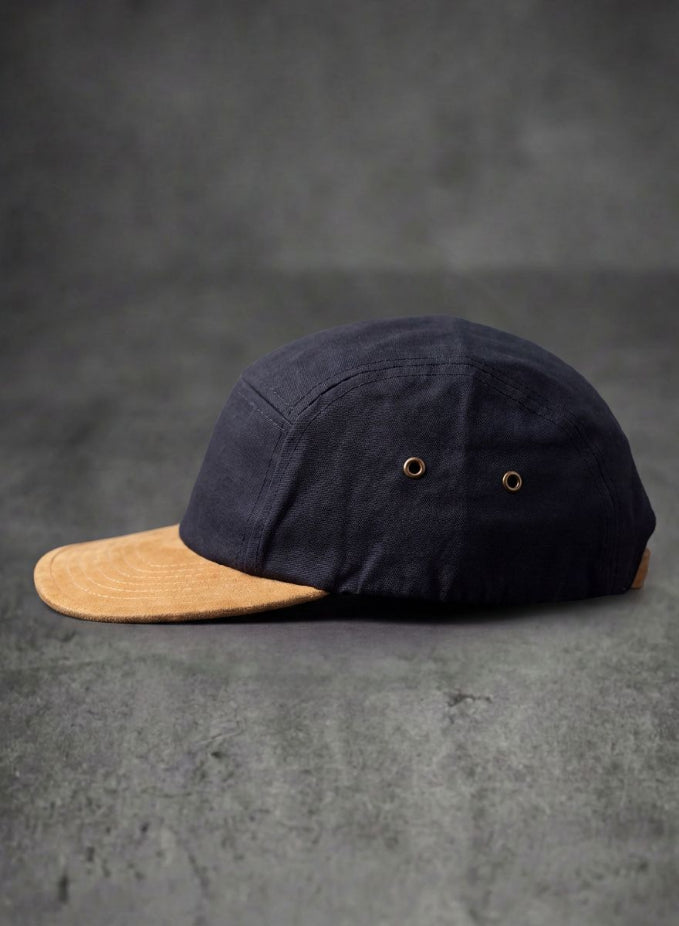 A dark black canvas baseball cap viewed from the back, featuring a brown leather strap and brass buckle closure, set against a textured gray background.