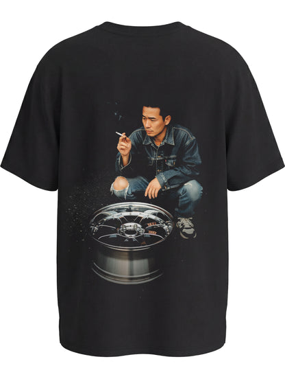 Black premium tshirt with a graphic of a japanese drifter person crouched over an alloy wheel expressing japanese drift culture on the back, on a white background