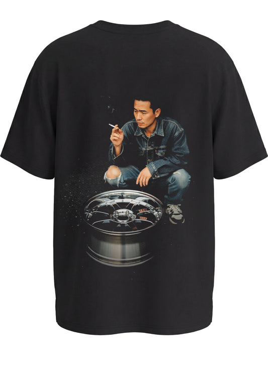 Black premium tshirt with a graphic of a japanese drifter person crouched over an alloy wheel expressing japanese drift culture on the back, on a white background