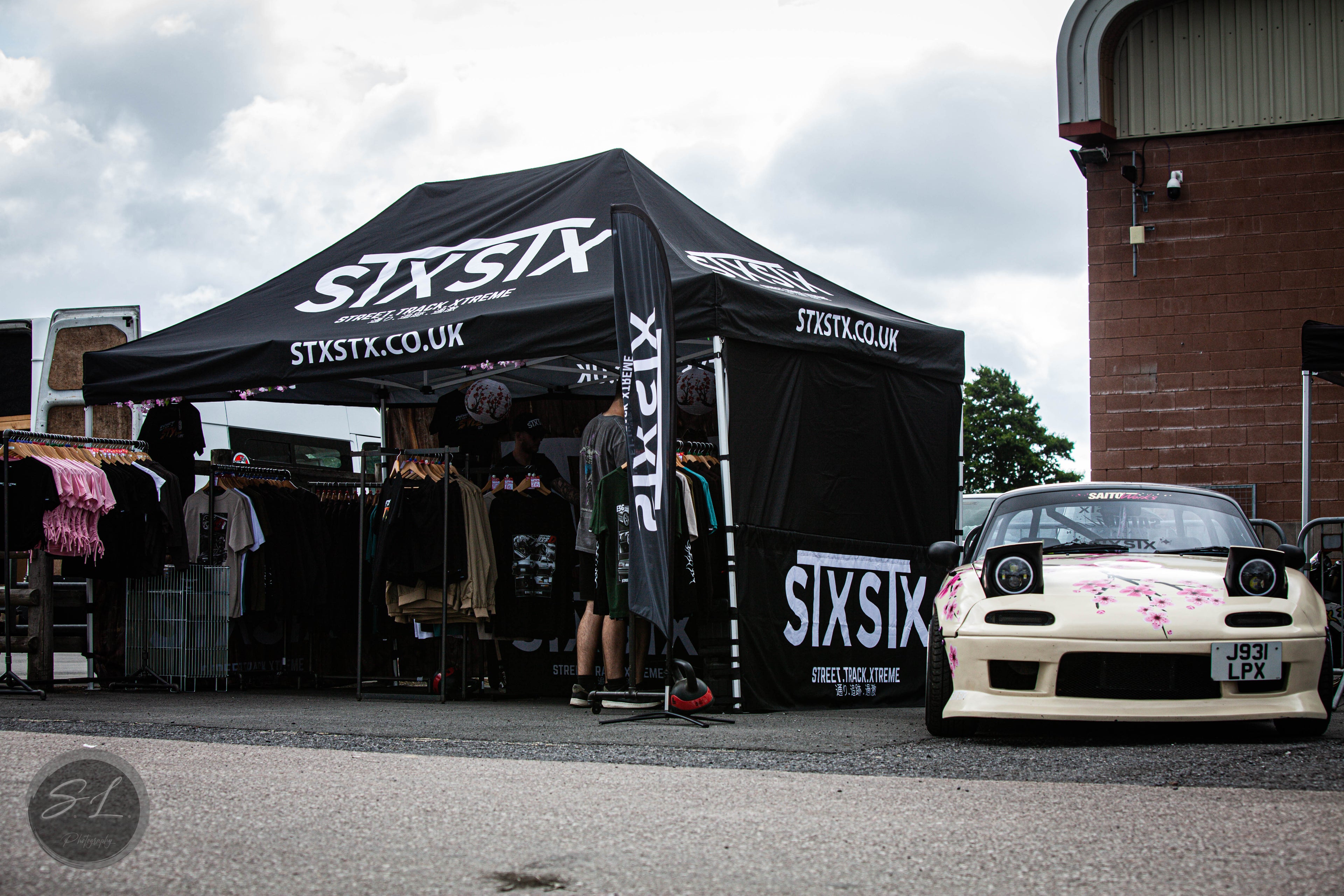 black gazebo with a shop at a carshow selling drift inspired merchandise with a mazda mx5 parked on display as a show car